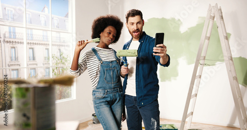 Joyful mixed-races young family couple wife and husband smiling posing with brushes taking selfie photos on smartphone in room. Home repair concept. Man and woman taking pictures during renovation