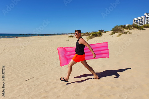 Young caucasian man with bright pink swimming mattress walking to seashore. Lifestyle