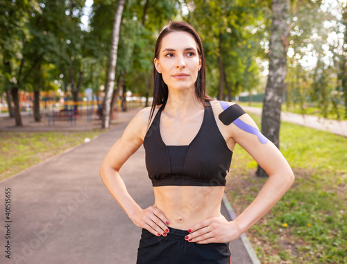 Fototapeta Naklejka Na Ścianę i Meble -  Portrait of attractive muscular brunette woman wearing black sports outfit, looking at camera. Young smiling female athlete posing with hands on waist, colorful kinesiotaping on body.