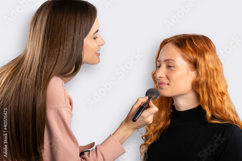 Makeup artist working at a makeup for a girl with red curly hair. White background. Beauty concept.