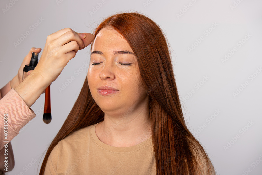 Fototapeta premium Makeup for a young girl. The master covers the foundation with a cosmetic sponge. White background.