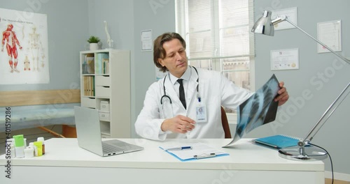 Portrait of busy Caucasian middle-aged man doctor in medical coat sitting at esk in hospital room, working on laptop, looking at X-ray scan results. coronavirus pandemic. treatment concept photo