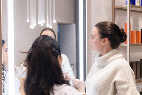 Two women, a beautician doctor and a client, stand at the mirror, at a consultation, discussing the upcoming procedures. Beautician talks about sculpting the face
