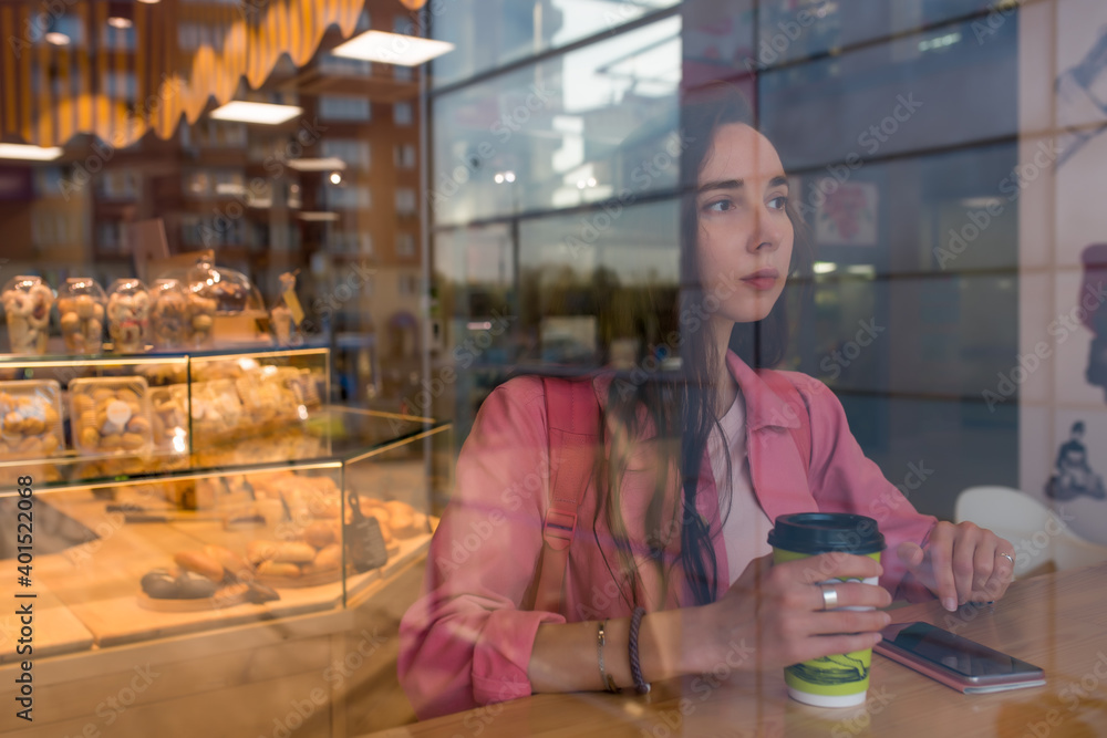 woman in summer city sits bakery cafe, holding cup coffee tea hand, resting table phone having lunch break breakfast. Free space copy of text. Reflection mirror. Casual clothes pink jacket, backpack.