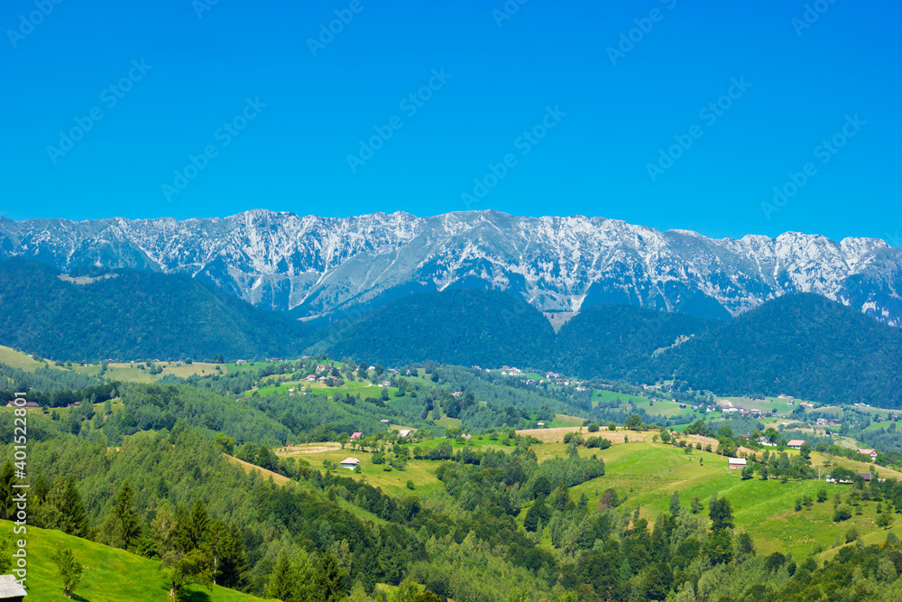 Stunning alpine landscape with green fields and high snowy Piatra Craiului mountains near Brasov. Panoramic view of mountain farms with houses. Bran, Transylvania, Romania, Europe. 