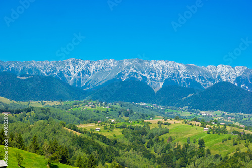 Stunning alpine landscape with green fields and high snowy Piatra Craiului mountains near Brasov. Panoramic view of mountain farms with houses. Bran  Transylvania  Romania  Europe. 