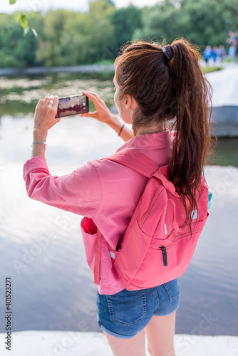 woman in summer in city near lake, river pond, photographs landscape on smartphone, view from back, a pink backpack behind her back, recording a video on Internet, application smartphone camera.