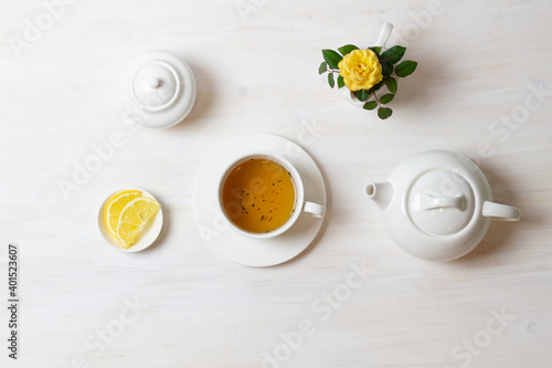 tea in cup with saucer, teapot, sugar bowl, yellow rose in milk jug and lemon on white background
