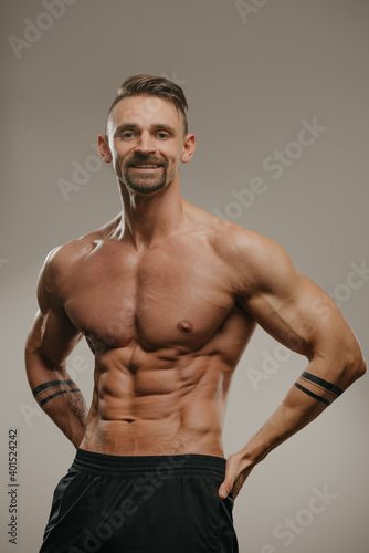 A smiling muscular man with a beard is posing. The athletic guy is squeezing his muscles. A bodybuilder with tattoos on his forearms is standing with his hands on his hips © Roman Tyukin