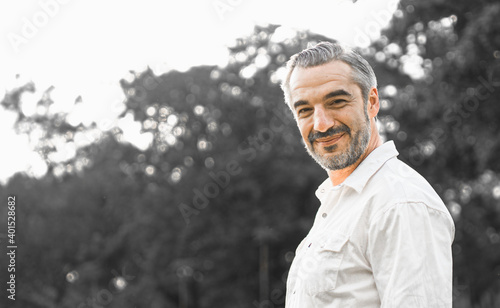 Portrait shot of happy, successful, attractive bearded middle-aged man smiling with a confident look toward the camera at a blur park background with copy space in natural sunlight.