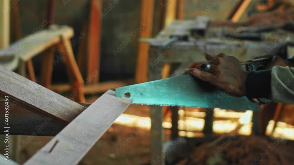 Old master with wrinkled hands saws a wooden board with a hand saw on wood in the workshop Old master with wrinkled hands saws a wooden board with a hand saw on wood in the workshop