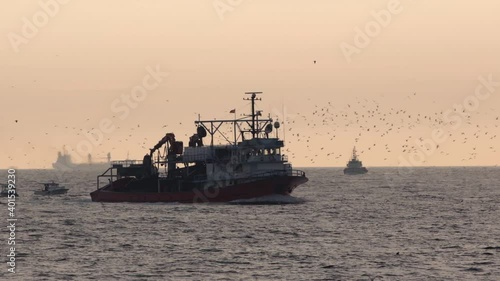 A trawler or fishing boat in the sea is coming back from fishing with many seagulls at sky.