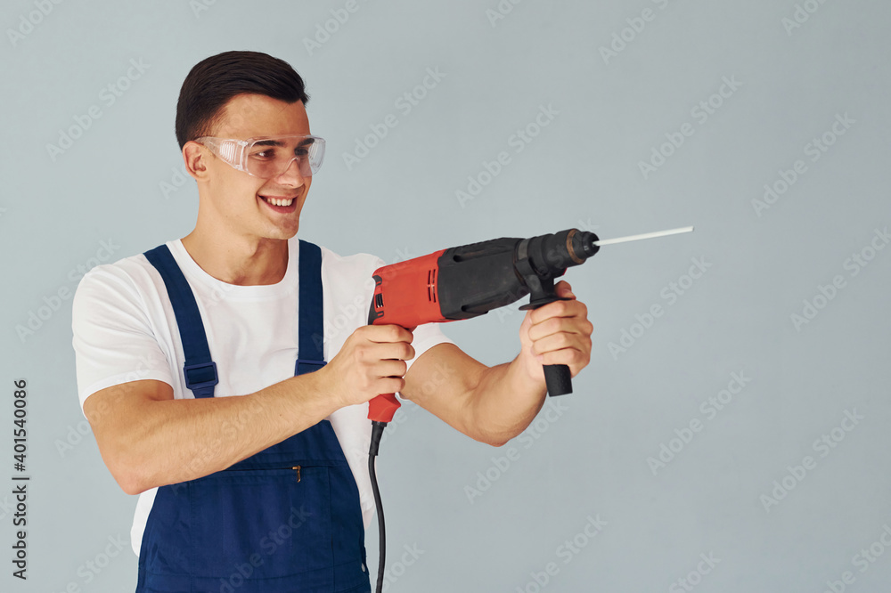 In protective eyewear and with drill in hands. Male worker in blue uniform standing inside of studio against white background In protective eyewear and with drill in hands. Male worker in blue uniform standing inside of studio against white background