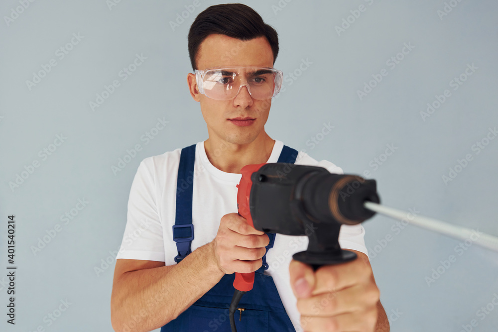 In protective eyewear and with drill in hands. Male worker in blue uniform standing inside of studio against white background In protective eyewear and with drill in hands. Male worker in blue uniform standing inside of studio against white background