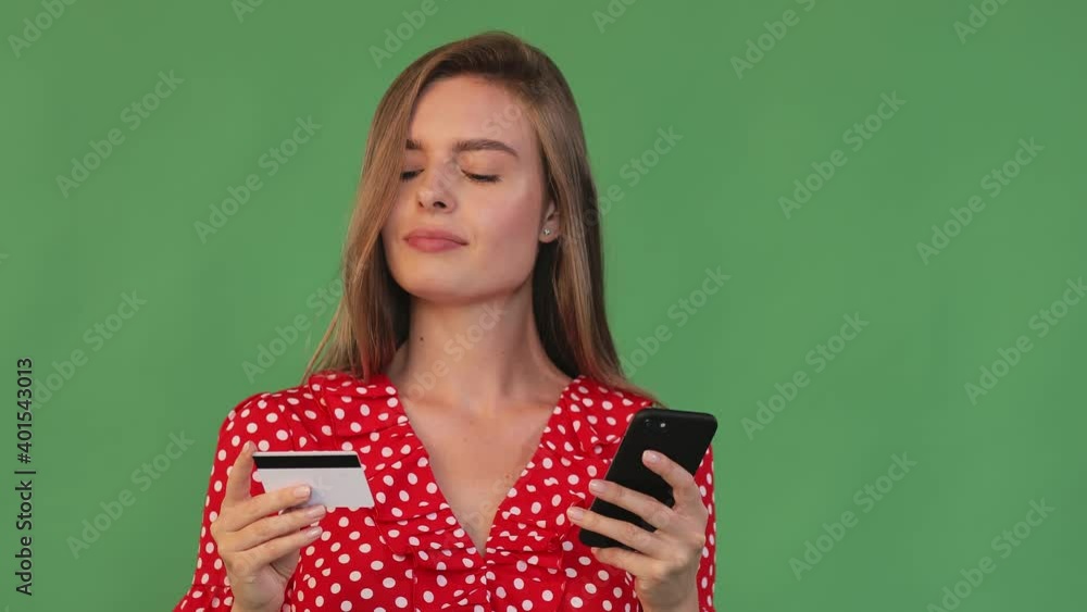 A girl holds a bank credit card and a phone in her hands, stands thoughtfully and hesitates to make purchases in an online store. A girl holds a bank credit card and a phone in her hands, stands thoughtfully and hesitates to make purchases in an online store.