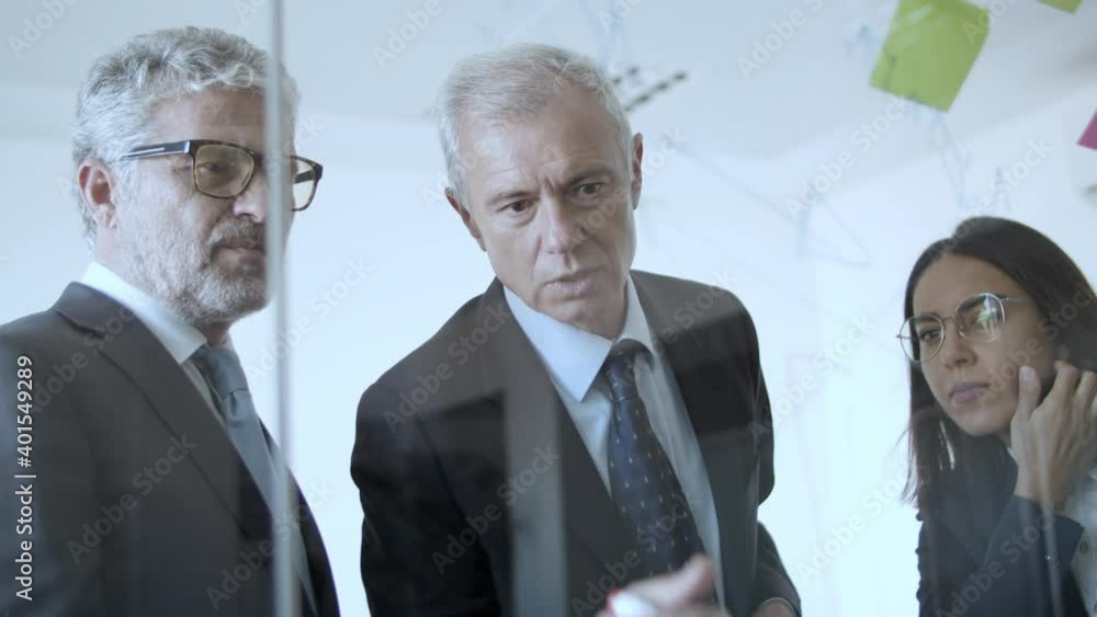 Team of different aged company managers working on project strategy, drawing charts on glass board with sticky notes. View through glass wall. Business plan concept Team of different aged company managers working on project strategy, drawing charts on glass board with sticky notes. View through glass wall. Business plan concept