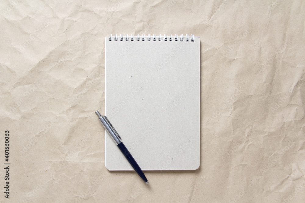 Gray notepad with white coiled spring and pen on a background of beige crumpled craft paper Gray notepad with white coiled spring and pen on a background of beige crumpled craft paper