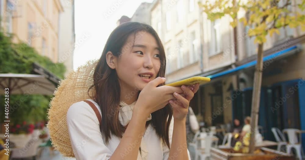 Close up portrait of happy young female tourist with hat standing on street and talking into speakerphone. Cheerful smiling Asian woman using internet virtual assistant app on cellphone outdoor.