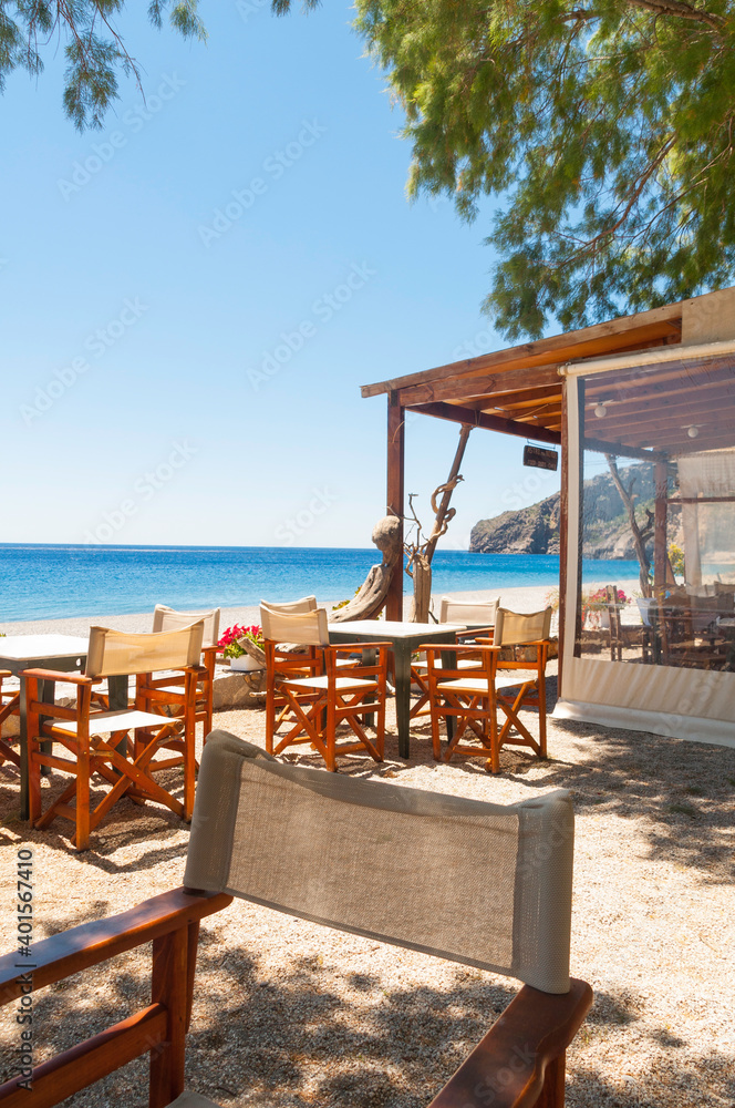 Cozy beach bar in a small secret bay on the north coast of the Greek island of Crete. Nice fabric armchairs and wooden tables stand in the sand. The sea can be seen in the background under a blue sky.