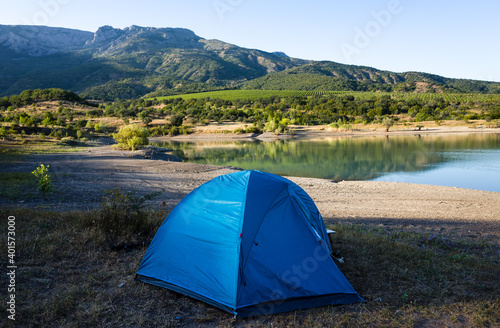 A blue tourist tent on the shore of a mountain lake against the backdrop of inaccessible rocks surrounded by a green forest.
