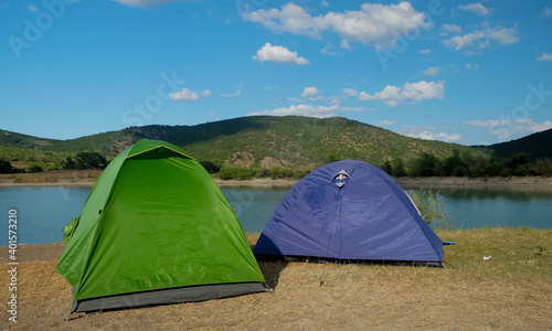 Multicolored tents on the shore of a mountain lake against the backdrop of inaccessible rocks surrounded by a green forest.