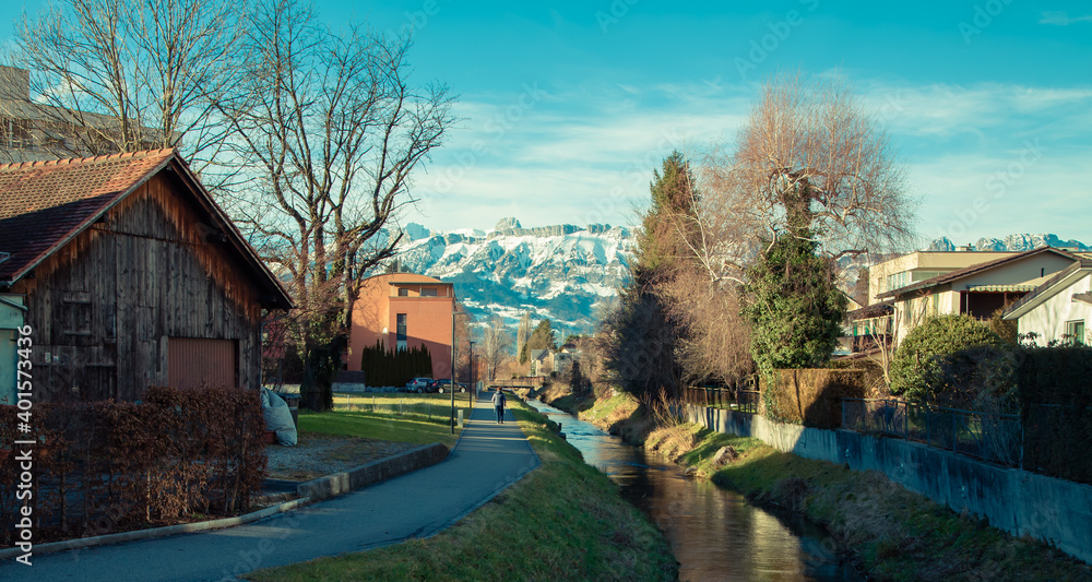 Alps mountains in Liechtenstein. Medieval Red House, calm narrow mountain river and jogging track, on the background of residential buildings, blue sky and snow-capped mountains. Liechtenstein, Vaduz Alps mountains in Liechtenstein. Medieval Red House, calm narrow mountain river and jogging track, on the background of residential buildings, blue sky and snow-capped mountains. Liechtenstein, Vaduz