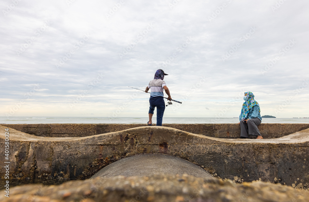 Port Dickson, Malaysia - December 15, 2020: Young man fishing on a breakwater on the beach. His wife with head scarf is sitting next to him and watching him. Fisherman throws his fishing rod 