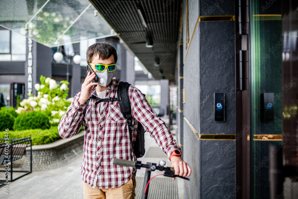 Fototapeta premium Courier young cute man using electric scooter delivered order to client on time, talking cell phone and waiting near front door of office, face mask protect by smog and car exhaust, delivery in city