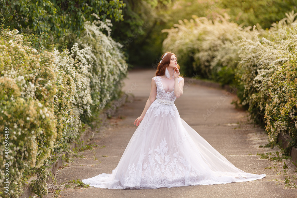 Beautiful redhead Bride in fantastic wedding dress in blooming garden. Growth Portrait in sunset light. Pretty young caucasian redhead girl walks in a garden and plays with her dress. Young princess Beautiful redhead Bride in fantastic wedding dress in blooming garden. Growth Portrait in sunset light. Pretty young caucasian redhead girl walks in a garden and plays with her dress. Young princess