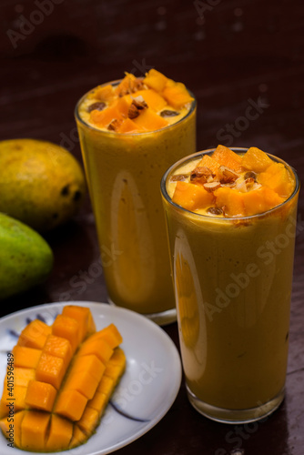 Mango lassi decorated with mango pulp raisin almonds and ripe mango in background mango slice in foreground