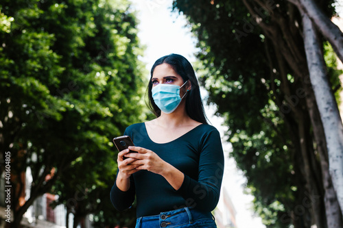 hispanic woman with facemask holding a phone and texting while she is walking in the street of a Latin America city photo