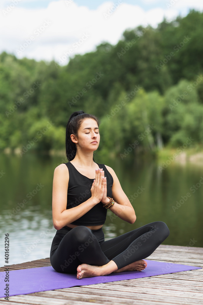 Young brunette woman meditates in lotus position on a wooden bridge by the river