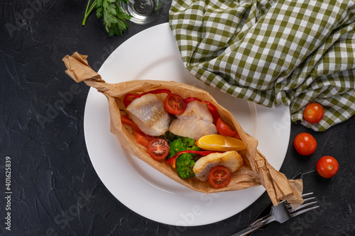 baked fish slices with vegetables in parchment on a white plate and dark background