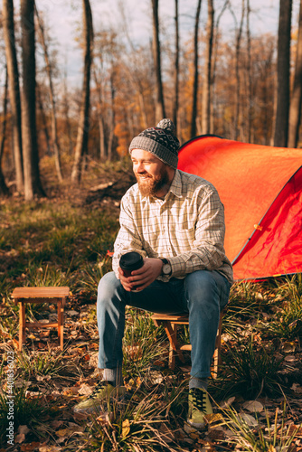 Full body delighted bearded man with takeaway cup smiling and looking away while sitting on stool near tent on campsite on autumn day in forest