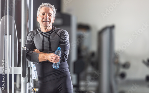 Aged man leans against a workout machine in a gym with a bottle of water in his hands