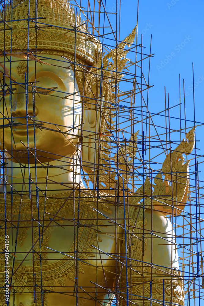 Close up image of large golden Buddha statue with scaffolding structure in under construction against blue sky at Thailand in vertical frame