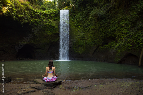 Young Caucasian woman meditating  practicing yoga at waterfall. Lotus pose. Hands in gyan mudra. Tibumana waterfall  Bali  Indonesia. View from back.