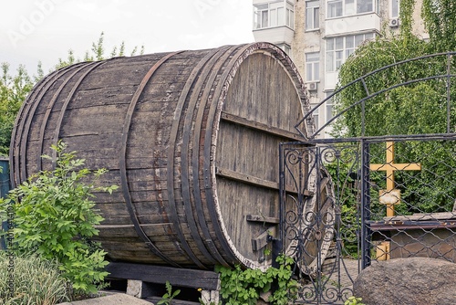 one large gray old wooden barrel stands outside in green vegetation by a black metal fence