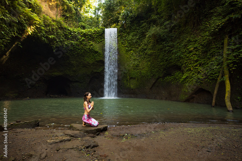 Young Caucasian woman sitting on the stone near the waterfall in Vajrasana or Diamond pose. Hands in namaste mudra. Tropical nature landscape. Yoga and pranayama concept. Tibumana waterfall  Bali