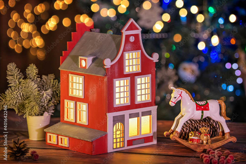 festive composition of a lamp in form of an old European house, toy horse, fir branches and cones against the background of defocused garlands
