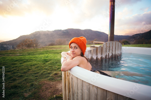 Fototapeta Naklejka Na Ścianę i Meble -  Beautiful young woman lay inside warm hot tub, heated by wood fire. Luxury outdoor vacation, natural hot spa or sauna resort. Woman relax in warm outdoor bath. Look in the camera and smile