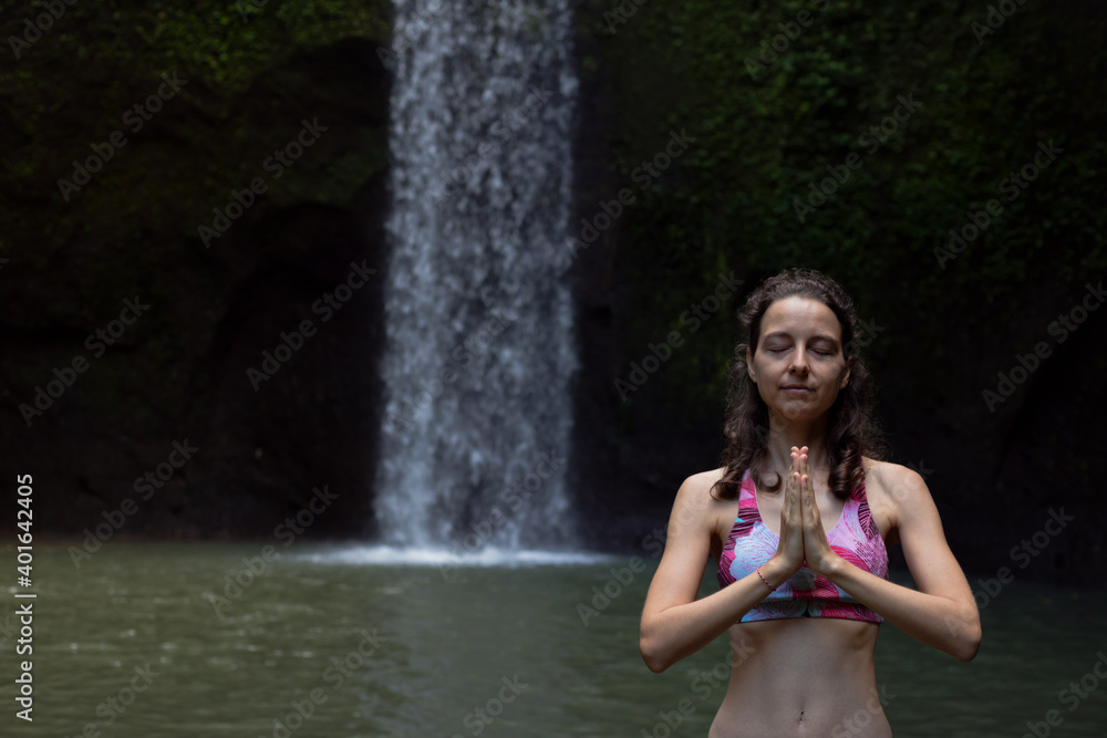 Fototapeta premium Close up of namaste mudra. Woman meditating, practicing yoga and pranayama with namaste mudra near waterfall. Yoga outdoor concept. Tibumana waterfall, Bali. Copy space.
