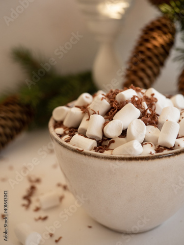 A cup of cocoa with marshmallows on the background of fir branches with cones in a vase and a burning candle on a light background in the morning light