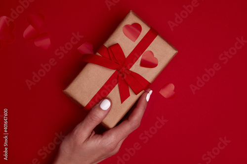Fototapeta Naklejka Na Ścianę i Meble -  Valentine day composition with copy space. Woman hand holding present or gift box wraped in craft paper with ribbon bow and red hearts confetti. Close up, top view, copy space