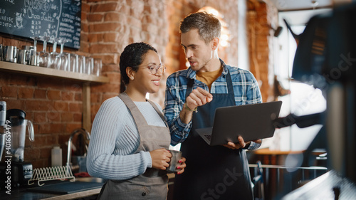Two Diverse Entrepreneurs Have a Team Meeting in Their Stylish Coffee Shop. Barista and Cafe Owner Discuss Work Schedule and Menu on Laptop Computer. Multiethnic Female and Male Restaurant Employees.