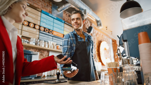 Female Customer Pays for Take Away Coffee with Contactless NFC Payment Technology on Smartphone to a Handsome Barista in Checkered Shirt in Cafe. Customer Uses Mobile to Pay Through Bank Terminal.