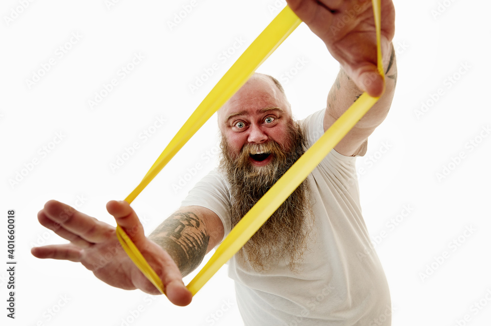 A laughing overweigh man stretches a yellow fitness elastic band and looks through it. Portrait isolated on a white background A laughing overweigh man stretches a yellow fitness elastic band and looks through it. Portrait isolated on a white background