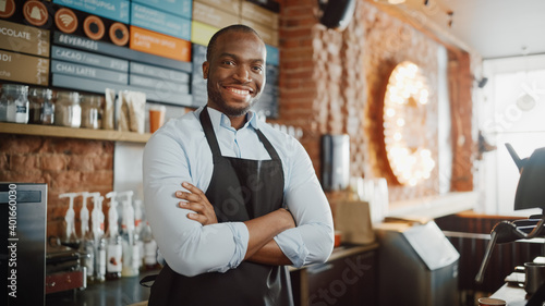 Handsome Black African American Barista with Short Hair and Beard Wearing Apron is Smiling in Coffee Shop Restaurant. Portrait of Happy Employee Behind Cozy Loft-Style Cafe Counter.