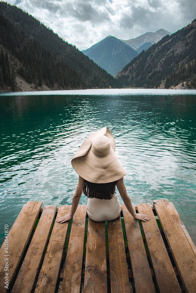 A girl in a hat sits on the pier and looks at the Kolsai lake - obrazy, fototapety, plakaty 