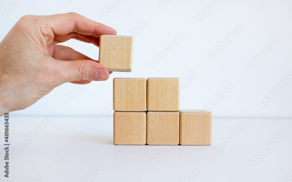 Symbol of building success foundation. Men hand put wooden cube on the pyramid from wooden blocks. Beautiful white background, copy space. Business and building success foundation concept. Symbol of building success foundation. Men hand put wooden cube on the pyramid from wooden blocks. Beautiful white background, copy space. Business and building success foundation concept.